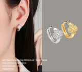 Close-up of a woman's ear wearing a silver hoop earring with gold accents, next to gold and silver clover-shaped earrings on a gray background.