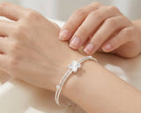 A delicate silver bangle bracelet with a central four-leaf clover charm and small clear stones, shown worn on a woman's wrist. The image features a soft-focus background with the model's hands and arms visible.