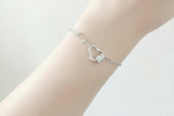 A silver chain bracelet with an open heart pendant and a small clear gemstone, shown worn on a person's wrist against a soft pink background. The text 'SILVER Factory Os.