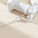 A silver heart-shaped pendant necklace with a textured center and a chain link detail, displayed on a soft pink background next to a white classical column prop. The brand name 'SILVER Factory Os' is visible in the top right corner.